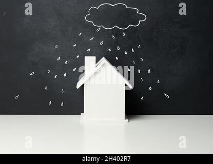 Holzhaus und eine gezeichnete Wolke mit Regen mit weißer Kreide auf einem schwarzen Kreidetafel. Konzept der Immobilienversicherung Stockfoto