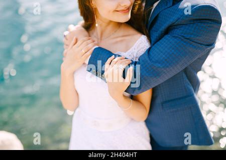 Der Bräutigam umarmt die Braut von hinten an den Schultern und küsst die Stirn an der Küste, Nahaufnahme Stockfoto