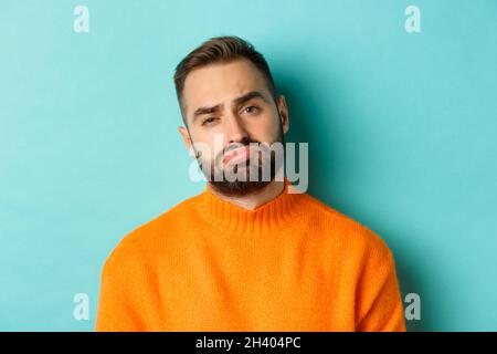 Nahaufnahme eines verärgerten jungen Mannes, der sich beschmutzt, enttäuscht und verzweifelt aussagt und in einem orangefarbenen Pullover gegen einen türkisfarbenen Hintergrund steht Stockfoto