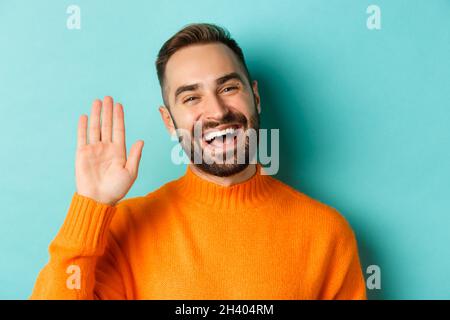 Nahaufnahme eines freundlichen jungen Mannes, der lächelt, mit der Hand winkt, um Hallo zu sagen, Sie begrüßt und auf hellblauem Hintergrund steht Stockfoto
