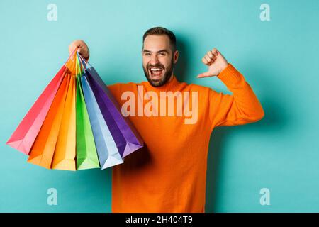 Hübscher bärtiger Mann in orangefarbenem Pullover, der Geschenke kauft, auf sich zeigt und Einkaufstaschen zeigt und über blauem Backgrou steht Stockfoto