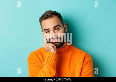 Nahaufnahme eines unamüsanten traurigen Mannes, der die Kamera ansah, das Gesicht an der Hand beugte und sich auf hellblauem Hintergrund auflehnte Stockfoto