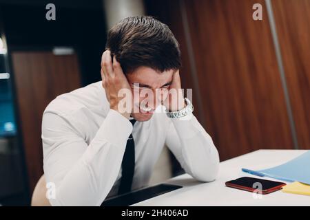 Trauriger junger Geschäftsmann sitzt und hält den Kopf mit den Händen am Bürotisch. Geschäftsleute Kopfschmerzen in der Krise und Depressionen danach Stockfoto