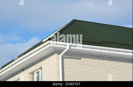 Ein Backsteinhaus mit einem grünen Metalldach, weißem Kunststoffsoffit und Faszien und einer Dachrinnenleitung, Downauslauf. Stockfoto