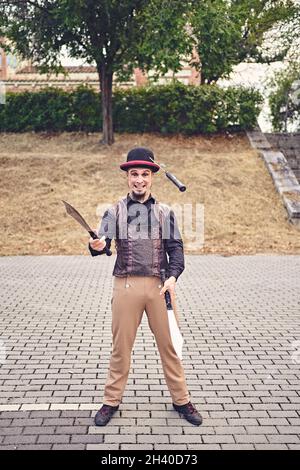 Full Body lustige männliche Darsteller in Kostüm lächelnd und jonglieren Messer, während auf dem Bürgersteig während der Show im Park stehen Stockfoto