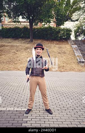 Full Body lustige männliche Darsteller in Kostüm lächelnd und jonglieren Messer, während auf dem Bürgersteig während der Show im Park stehen Stockfoto