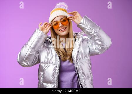 Stilvolle freche gewagte blonde europäische Mädchen handeln cool tragen stilvolle Sonnenbrille silberne Jacke Winterhut Überprüfung Rahmen Nase tilti Stockfoto