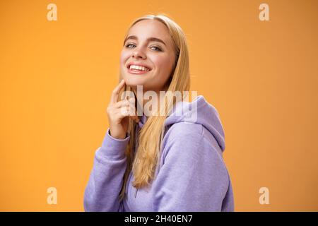 Attraktive freche selbstbewusste kaukasische blonde Frau in lila Hoodie Drehkamera berühren Kinn frech Lächeln Kamera Check out Stockfoto