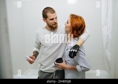 Lustiges 30-jähriges Paar, das sich im Badezimmer umarmt, Mann hält Zahnbürste. Er hat eine prothetische bionische Hand. Stockfoto