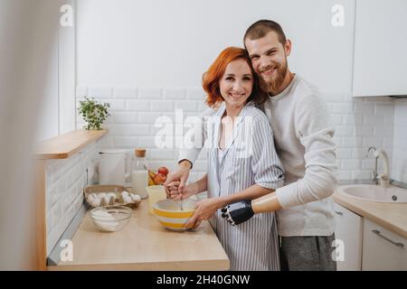 Porträt eines dreißigjährigen Paares, das in der Küche zusammen kocht und Eier brüstelt. Mann umarmt Frau von hinten. Er hat eine prothetische bionische Hand. Stockfoto