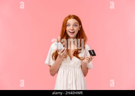 Beeindruckt und aufgeregt, glücklich Rotschopf Mädchen in weißem Kleid, erstaunt, wie einfach sie den Kauf gemacht, halten Smartphone und Kreditkarte, Stockfoto