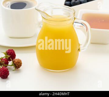 Frisch gepresster Saft in einer transparenten Glasdekanter, eine Tasse Kaffee, Honig auf einem weißen Tisch. Frühstück am Morgen Stockfoto