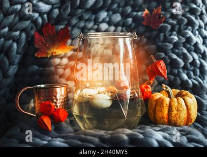 Stillleben im Herbst mit Glas mit Kerzenlicht, Teetasse, Kürbis und orangefarbenen Herbstblättern auf grauer Wolldecke. Gemütlicher Hygge-Lifestyle. Vorderansicht. Stockfoto