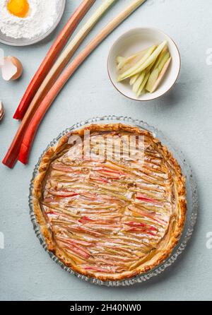 Hausgemachter Rhabarberkuchen auf grauem Küchentisch mit Rhabarber, Mehl mit Eigelb, Eierschale und Schüssel. Saisonales Backen zu Hause. Draufsicht. Stockfoto