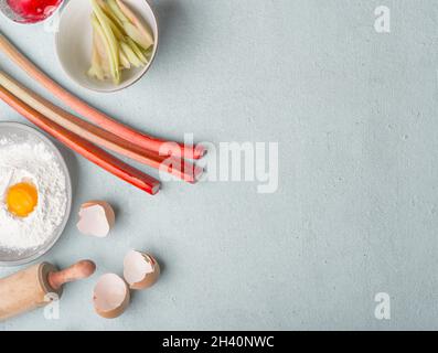 Lebensmittelhintergrund mit Rhabarber, Eigelb, Mehl, Eierschale, Holzrollholz, Geschirrtücher und Gabeln auf grauem Küchentisch. Kuchen Zubereitung zu Hause mit Stockfoto