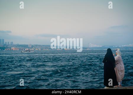 ISTANBUL, TÜRKEI - 17. JULI 2021: Zwei junge muslimische Frauen, die Hijabs tragen, fotografieren am Ufer des Bosporus mit der Stadt Stockfoto