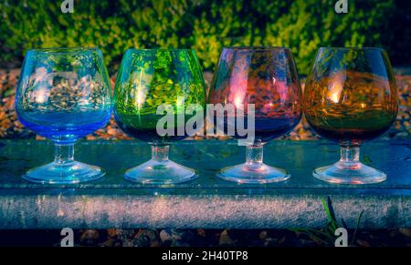 Eine Auswahl an farbenfrohen Schnaps- oder Likörgläsern in einer Reihe auf einer Granitplatte in einem üppigen Garten Stockfoto
