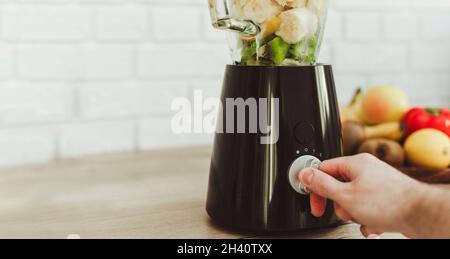 Mann Schalten Sie den stationären Mixer ein, um einen gesunden grünen Smoothie zuzubereiten. Zutaten auf dem Tisch in der Küche Stockfoto