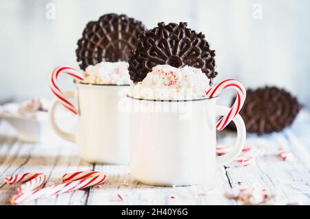 Zwei Tassen heißen Kakao mit Schlagsahne, zerdrückten Zuckerstöcken und Pizzelle Plätzchenwafern für Weihnachten. Selektiver Fokus mit verschwommenem Vordergrund. Stockfoto