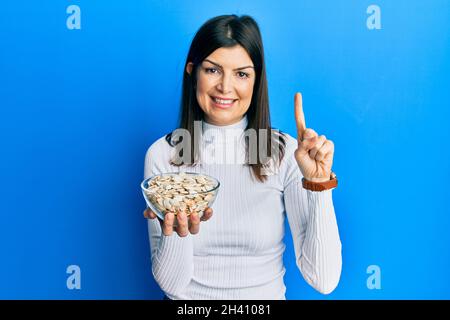 Junge hispanische Frau hält Kürbiskerne Schüssel lächelnd mit einer Idee oder Frage zeigen Finger mit glücklichen Gesicht, Nummer eins Stockfoto