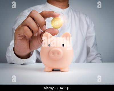 Einsparungs- und Investitionskonzept. Pinkes Sparschwein und die Hand eines Geschäftsmannes im weißen Hemd hält und zeigt eine glänzende goldene Geldmünze oder Bitcoin auf wh Stockfoto