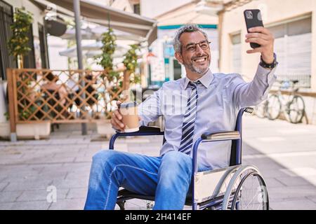 Ein Mann aus dem mittleren Alter, der auf dem Rollstuhl sitzt, macht Selfie mit dem Smartphone auf der Straße Stockfoto