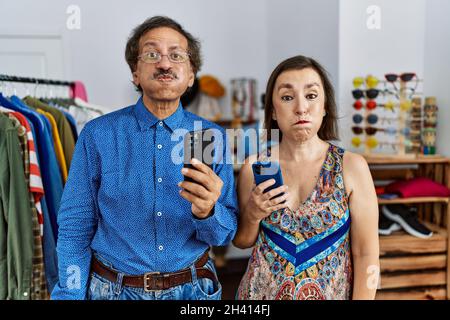 Interrassisches Paar im mittleren Alter im Einzelhandel mit Smartphone-Puffing Wangen mit lustigem Gesicht. Mund mit Luft aufgeblasen, verrückter Ausdruck. Stockfoto