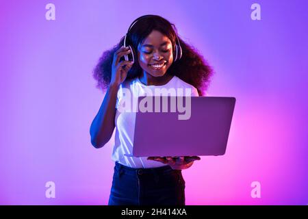 Junge schwarze Frau mit Videoanruf, mit Laptop-Computer, tragen drahtlose Kopfhörer in Neonlicht Stockfoto