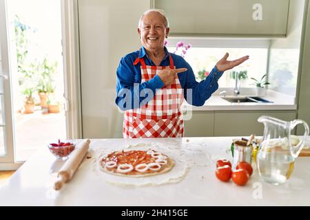 Älterer Mann mit grauen Haaren, der Pizza in der heimischen Küche kocht, staunt und lächelt zur Kamera, während er mit der Hand vorstellt und mit dem Finger zeigt. Stockfoto