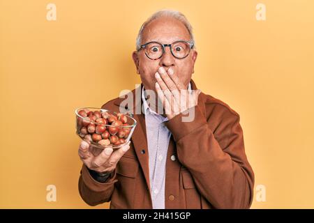 Älterer Mann mit grauem Haar, der Haselnüsse hält, die den Mund mit der Hand bedecken, schockiert und vor Fehlern fürchtet Stockfoto