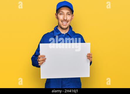Glatze Mann mit Bart tragen Baumeister Jumpsuit Uniform halten leere Banner sieht positiv und glücklich stehen und lächeln mit einem selbstbewussten Lächeln showi Stockfoto