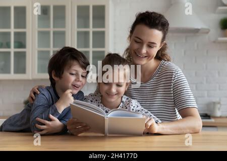 Lächelnde kleine Kinder lesen Buch mit fürsorglicher Mutter. Stockfoto