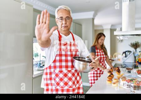 hispanische Paare mittleren Alters tragen Schürze Kochen hausgemachtes Gebäck mit offener Hand dabei Stop-Zeichen mit seriöse und selbstbewusste Ausdruck, Verteidigung Geste Stockfoto