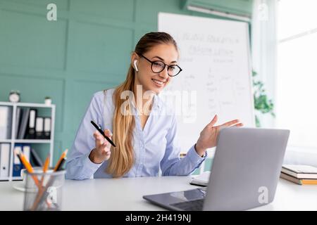 E-Learning. Die Lehrerin führt Videoanruf mit Schülern und der Klassengruppe, benutzt einen Laptop und trägt schnurlose Ohrstöpsel Stockfoto