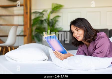 Eine Frau mittleren Alters ruht sich aus, während sie im Bett liegt und ein Buch liest. Das Konzept der Erholung von der Technik und dem Trubel der Großstadt. Stockfoto