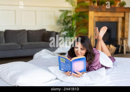 Eine Frau mittleren Alters ruht sich aus, während sie im Bett liegt, während sie ein Buch liest. Digitales Detox-Konzept. Stockfoto