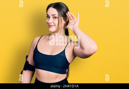 Junge hispanische Frau trägt Sportkleidung lächelnd mit der Hand über dem Ohr und hört Gerüchten oder Klatsch zu. Taubheitskonzept. Stockfoto