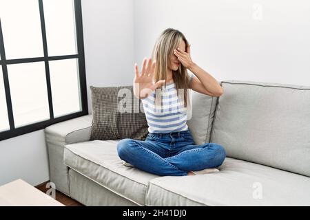 Asiatische junge Frau, die zu Hause auf dem Sofa sitzt und die Augen mit den Händen bedeckt und mit trauriger und angstvoller Geste stoppt Stockfoto