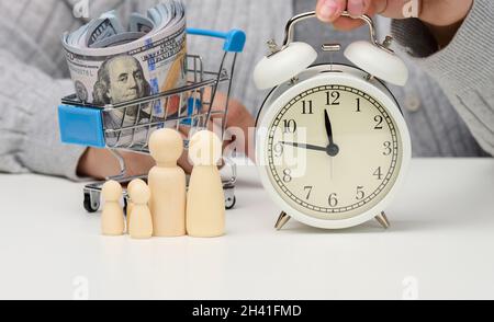 Holzfiguren einer Familie, eine weibliche Hand hält einen Wecker auf einem weißen Tisch. Konzept Zeit ist Geld, Budgetplanung. Zeit Stockfoto