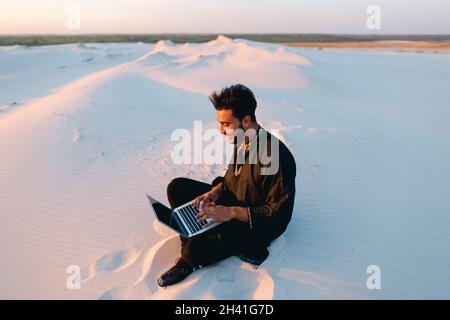 Ein hübscher junger arabischer Mann schreibt mit einem Laptop über das Internet und sitzt bei Sonnenuntergang auf Sand inmitten einer sandigen Wüste. Stockfoto