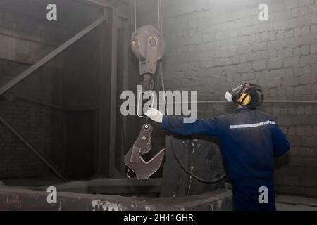 Ein Mann in einem Arbeitsanzug und einem Atemschutzgerät nimmt in der Werkshalle mit der Hand einen Haken und einen industriellen Hebemechanismus. Stockfoto