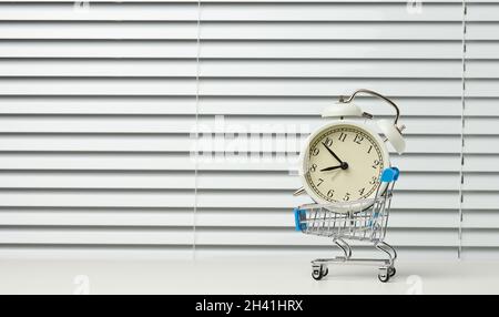 Mini-Einkaufswagen aus Metall auf Rädern und in der Mitte eines runden Weckers auf einem weißen Tisch. Beginn der Rabatte, Verkauf, Cop Stockfoto