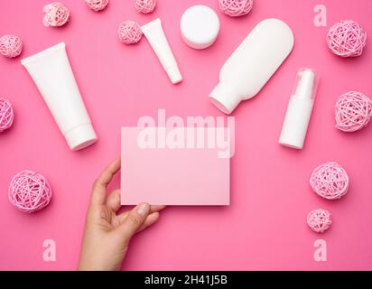 Weibliche Hand hält leeres rosa Papier auf einem rosa Hintergrund und Glas, Flasche und leere weiße Plastikröhrchen für Kosmetika auf einem rosa Stockfoto