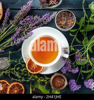Tee, quadratischer Schuss über dem Kopf. Kräuter, Blumen und Früchte um eine Tasse Tee Stockfoto