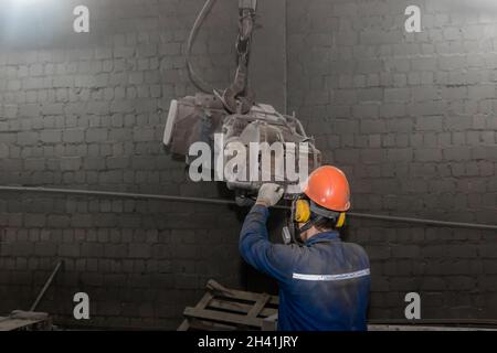 Ein Arbeiter in Overalls, Schutzhelm und Atemschutzmaske steuert schwere Schleifgeräte in der Werkstatt eines Industriewerks. Stockfoto