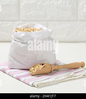 Roher Haferbrei in einem weißen Papierbeutel und ein Holzlöffel auf einem weißen Tisch, Frühstücksbrei Stockfoto