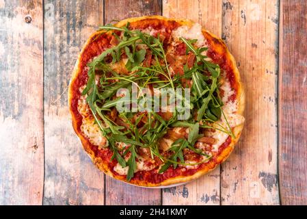 Burrata-Käse-Pizza mit Speckstücken, Tomaten, Mozzarella-Käse und Rucola-Blättern auf Holztisch Stockfoto