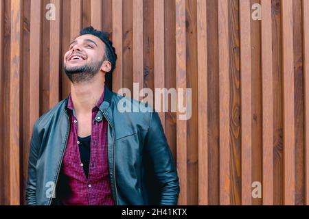 Nett aussehender arabischer junger Mann steht in der Nähe von Wall und Smiles, lacht, blickt auf Kamera und auf Seiten auf dem Hintergrund von Treppen im Freien. Stockfoto