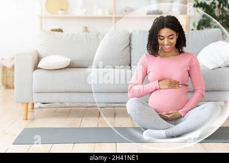 Glückliche ruhige junge afroamerikanische Schwangerin meditiert und berührt den Bauch im Wohnzimmer Stockfoto