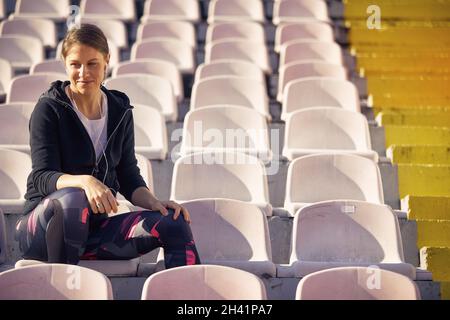 Schöne junge Erwachsene kaukasische Frau, die im Stadion mit Kopfhörern in den Ohren sitzt, lächelt und mit ihrem Handy spricht Stockfoto